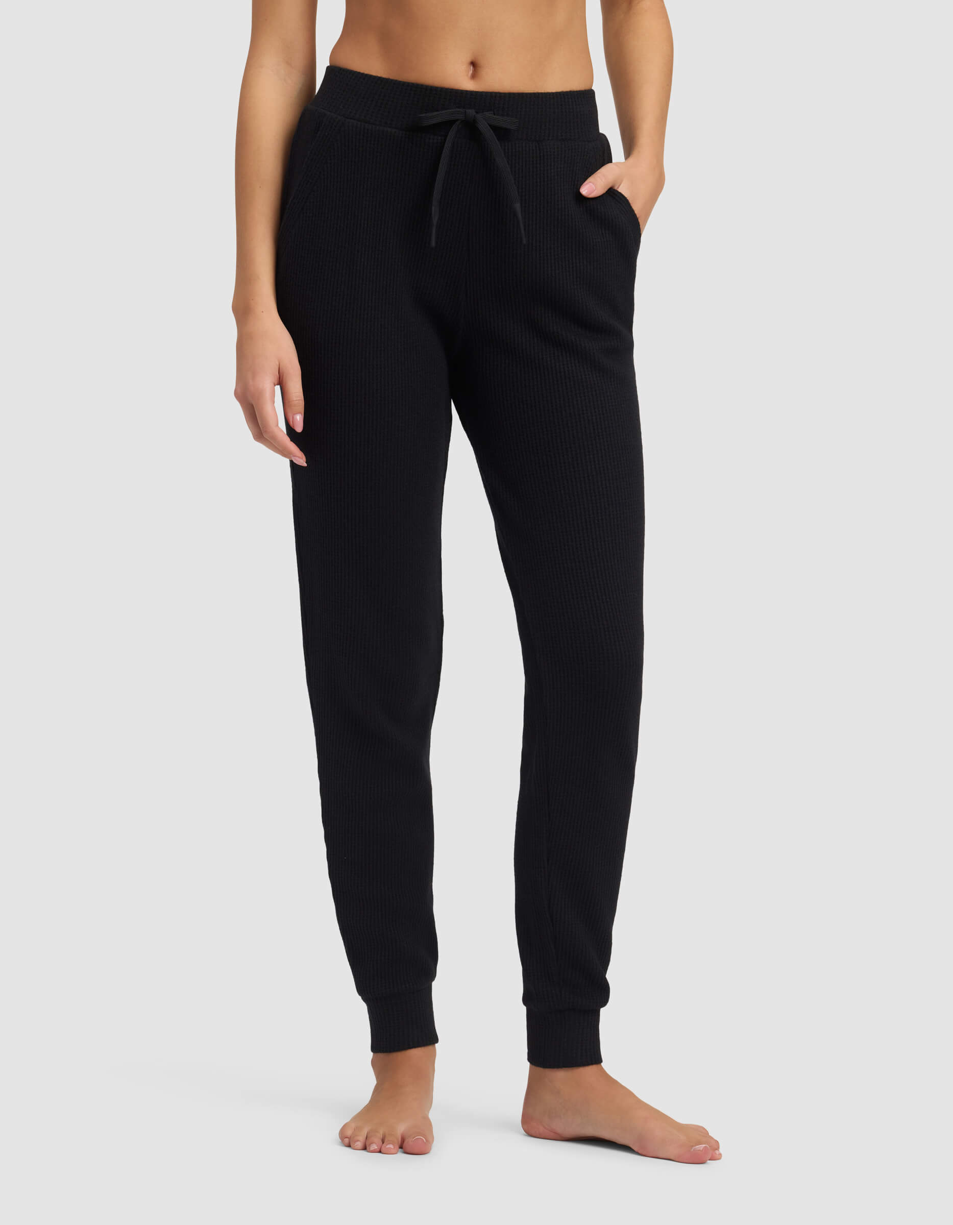 A person stands barefoot against a light gray background, modeling the lower torso and legs in Cozy Earth's Women's Blake Waffle Jogger, featuring a ribbed texture, drawstring waist, and pockets.