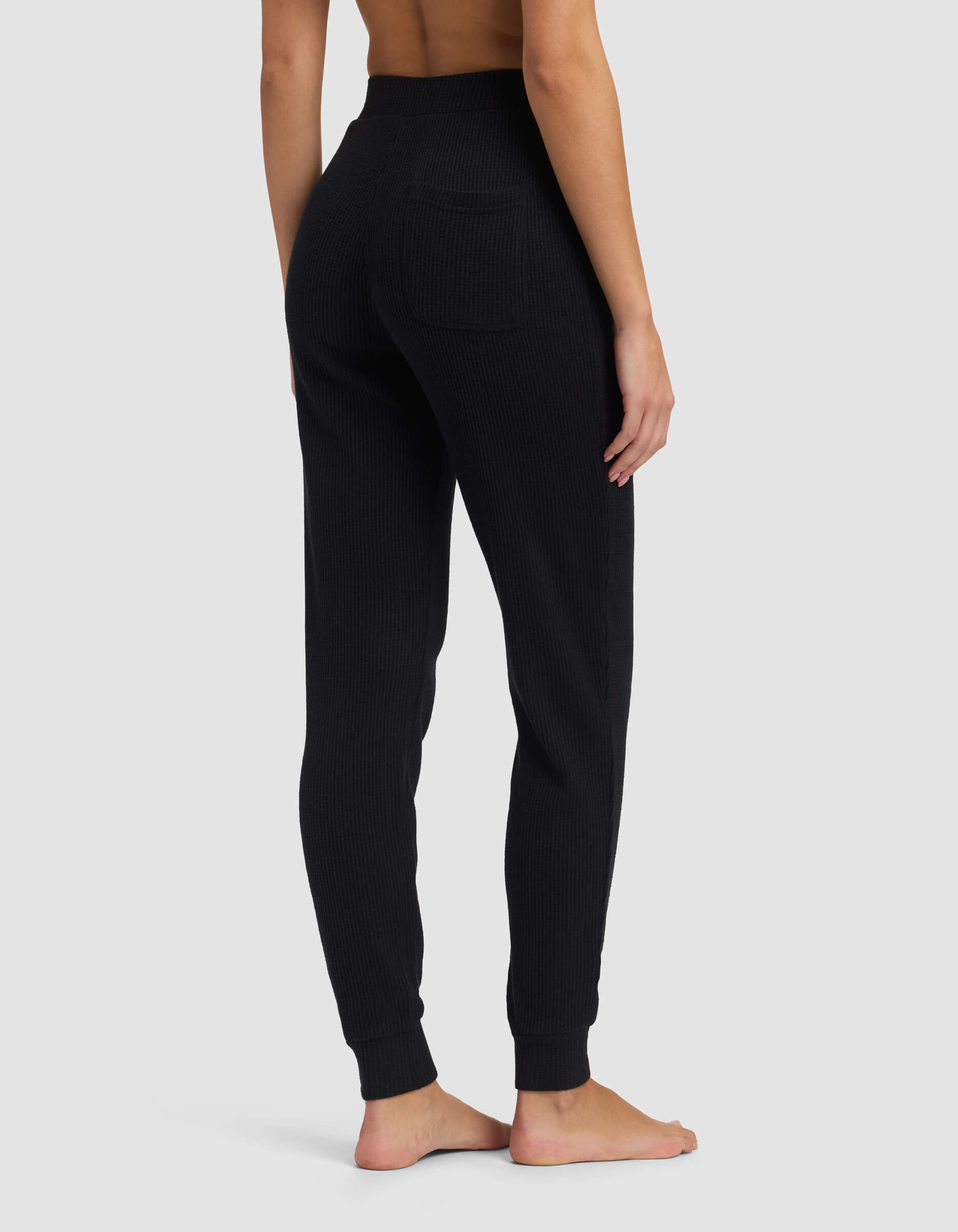 A person stands barefoot on a gray background, shown from the back, wearing Cozy Earth's Women's Blake Waffle Jogger in black. The high-waisted ribbed knit pants feature a single back pocket. The upper body is not fully visible.