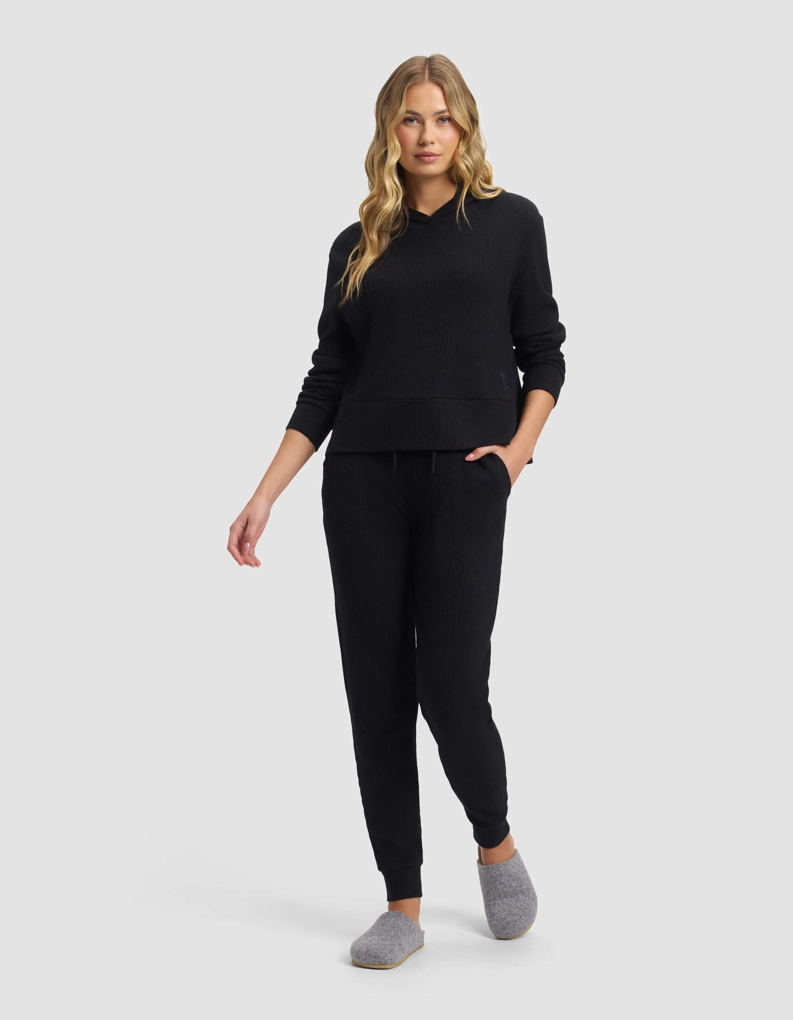 A woman with long blonde hair wears the Cozy Earth Women's Blake Waffle Jogger set with gray slippers, standing and looking forward against a plain light gray background.