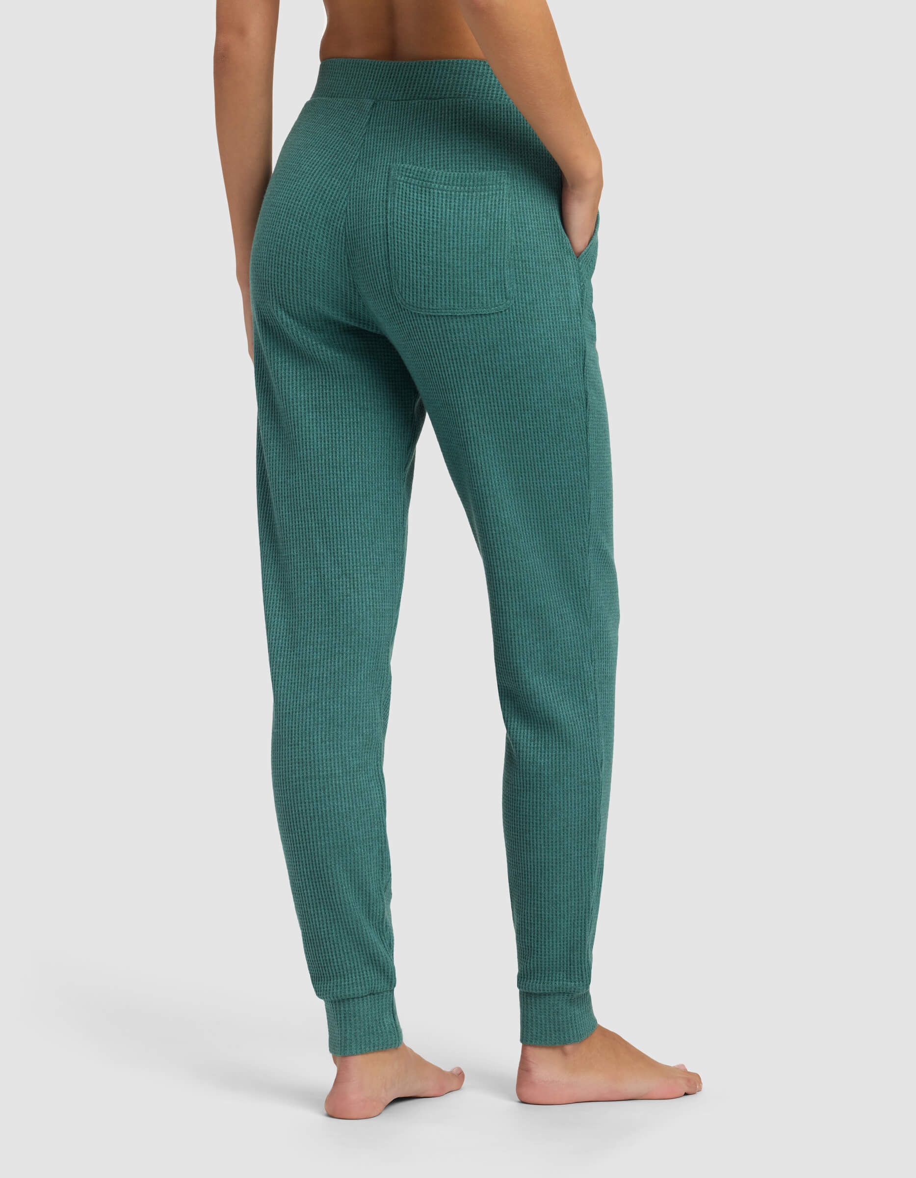 A person stands barefoot, facing away, wearing the Cozy Earth Women's Blake Waffle Jogger in teal with a right-side pocket, fitted waistband, and ankle cuffs. The background is plain light gray.|Color:Sea Green