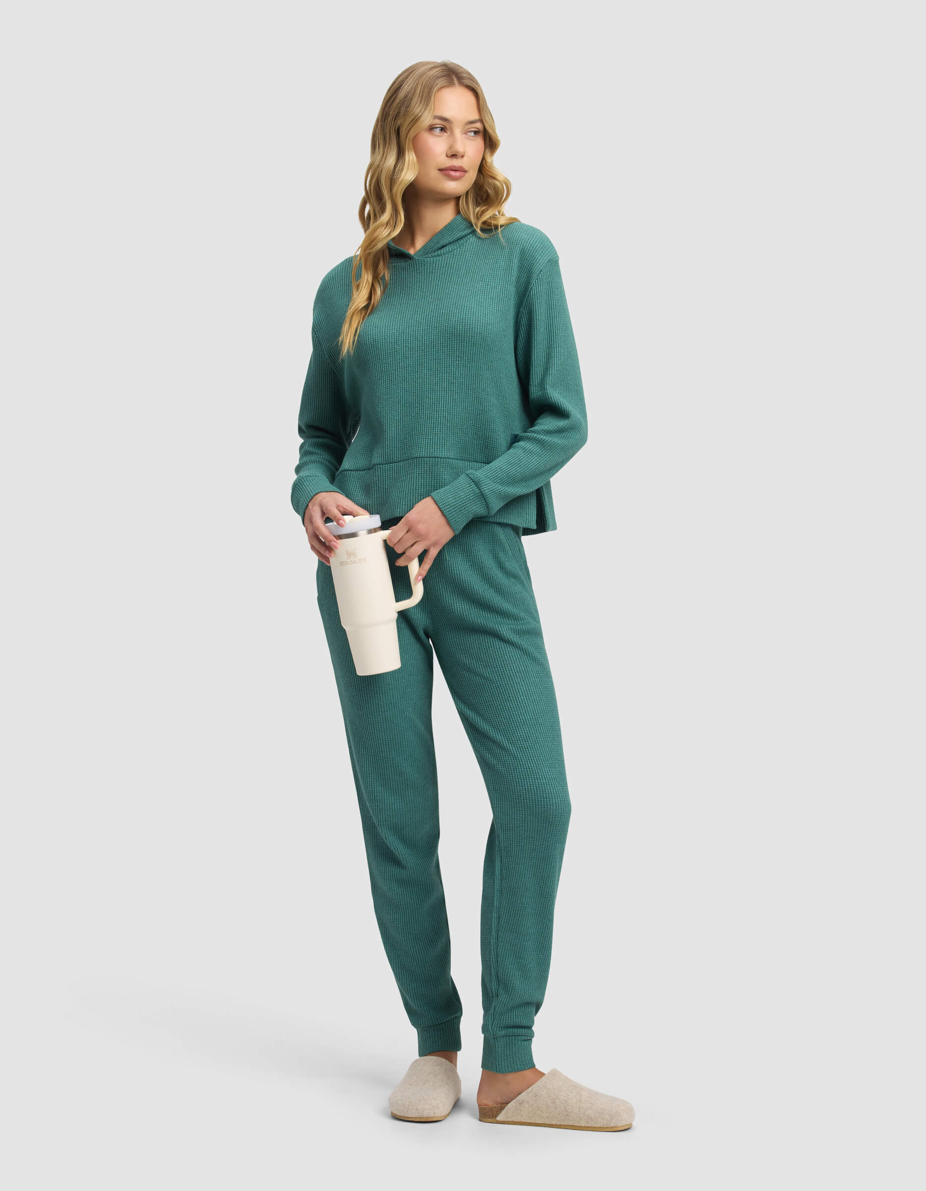 A woman wearing Cozy Earth's Women's Blake Waffle Jogger set in teal, with a hoodie and joggers, holds a white travel mug. She pairs the lounge set with beige slippers and stands against a plain light gray background.