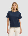 A woman with wavy blonde hair wears the Cozy Earth Women's All Day Tee in navy blue and light beige pants, standing against a light gray background and facing forward with a neutral expression.|Color:Eclipse