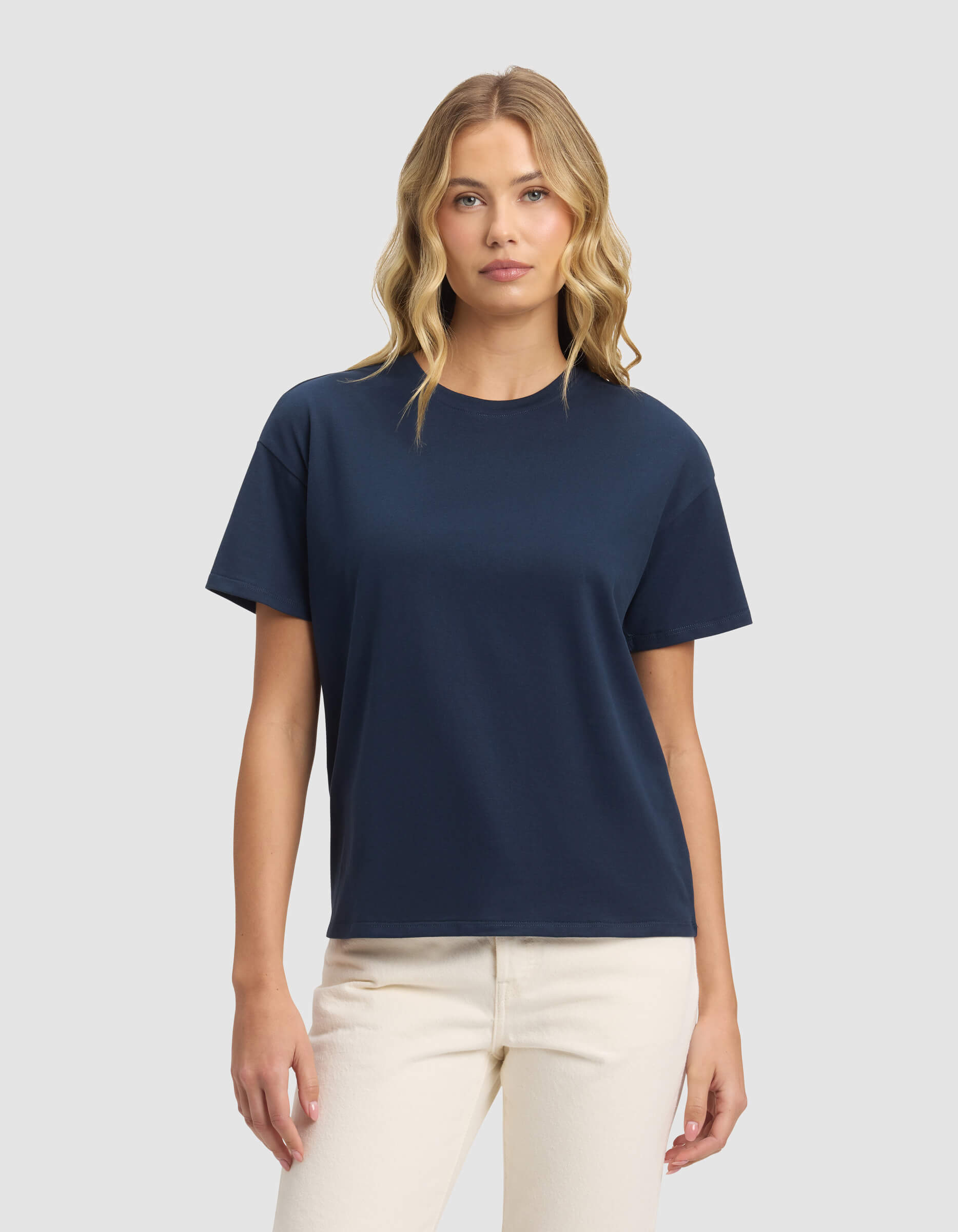 A woman with wavy blonde hair wears the Cozy Earth Women's All Day Tee in navy blue and light beige pants, standing against a light gray background and facing forward with a neutral expression.|Color:Eclipse
