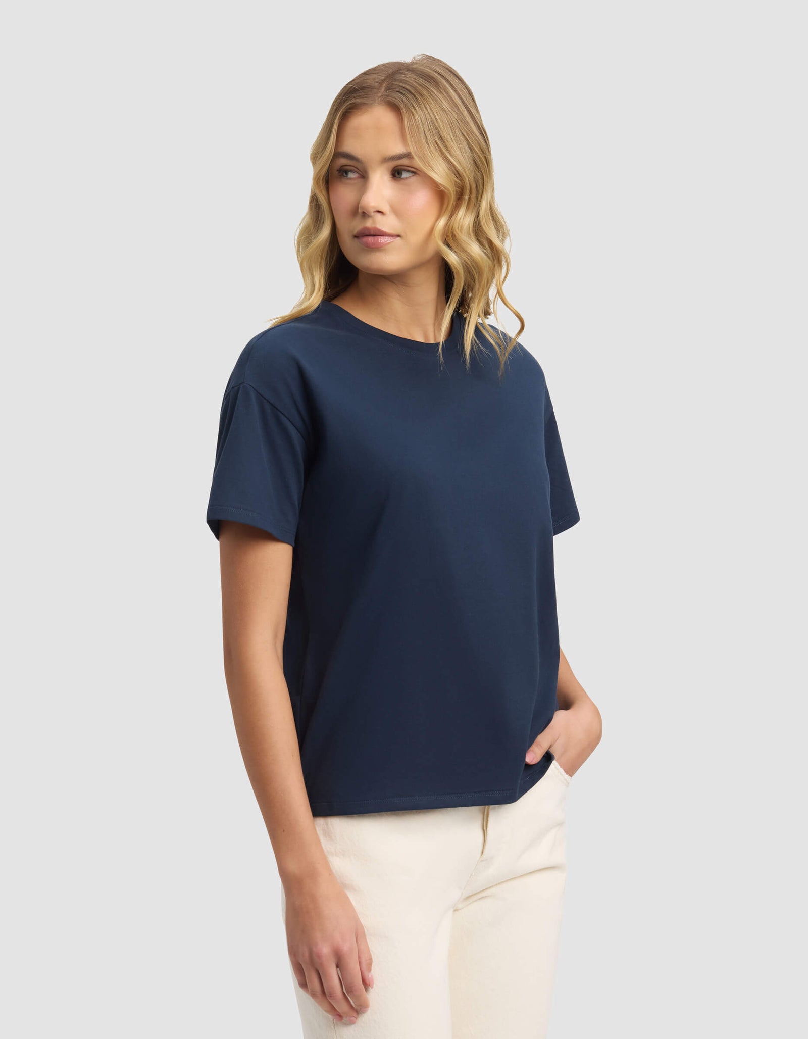 A woman with wavy blonde hair wears the Cozy Earth Women's All Day Tee in navy and off-white pants, standing against a plain light gray background, gazing slightly left with one hand in her pocket.