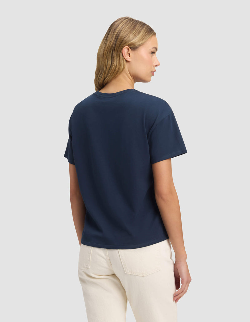 A woman with blonde hair models the Cozy Earth Women's All Day Tee in navy, paired with off-white pants, standing with her back to the camera against a plain light gray background.