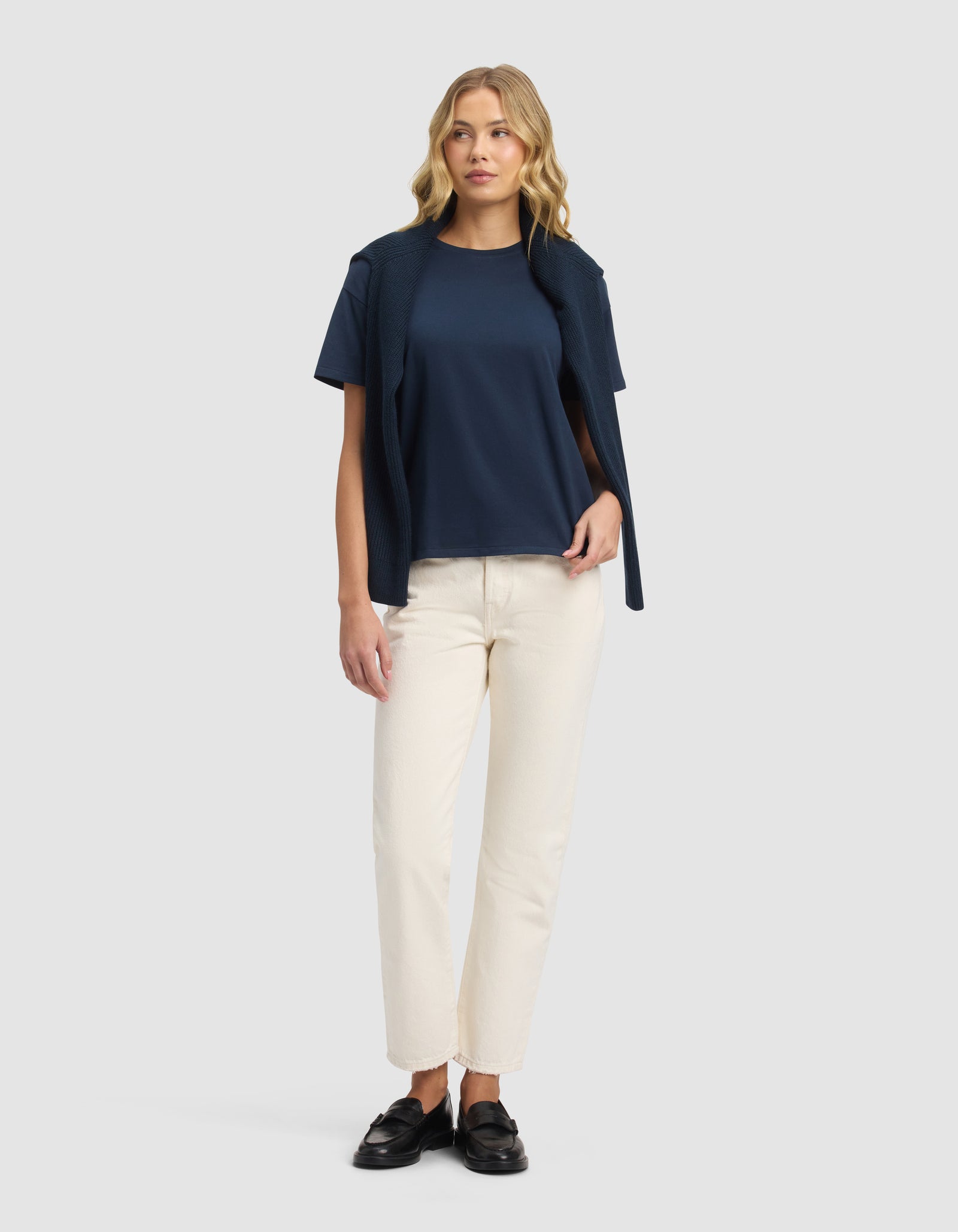 A woman with wavy blonde hair and a relaxed posture wears the Cozy Earth Women's All Day Tee in navy, drapes a matching sweater over her shoulders, pairs it with off-white straight-leg pants, and completes the look with black loafers.