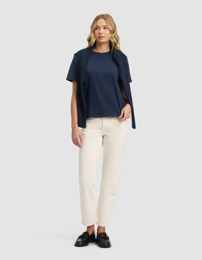 A woman with wavy blonde hair and a relaxed posture wears the Cozy Earth Women's All Day Tee in navy, drapes a matching sweater over her shoulders, pairs it with off-white straight-leg pants, and completes the look with black loafers.