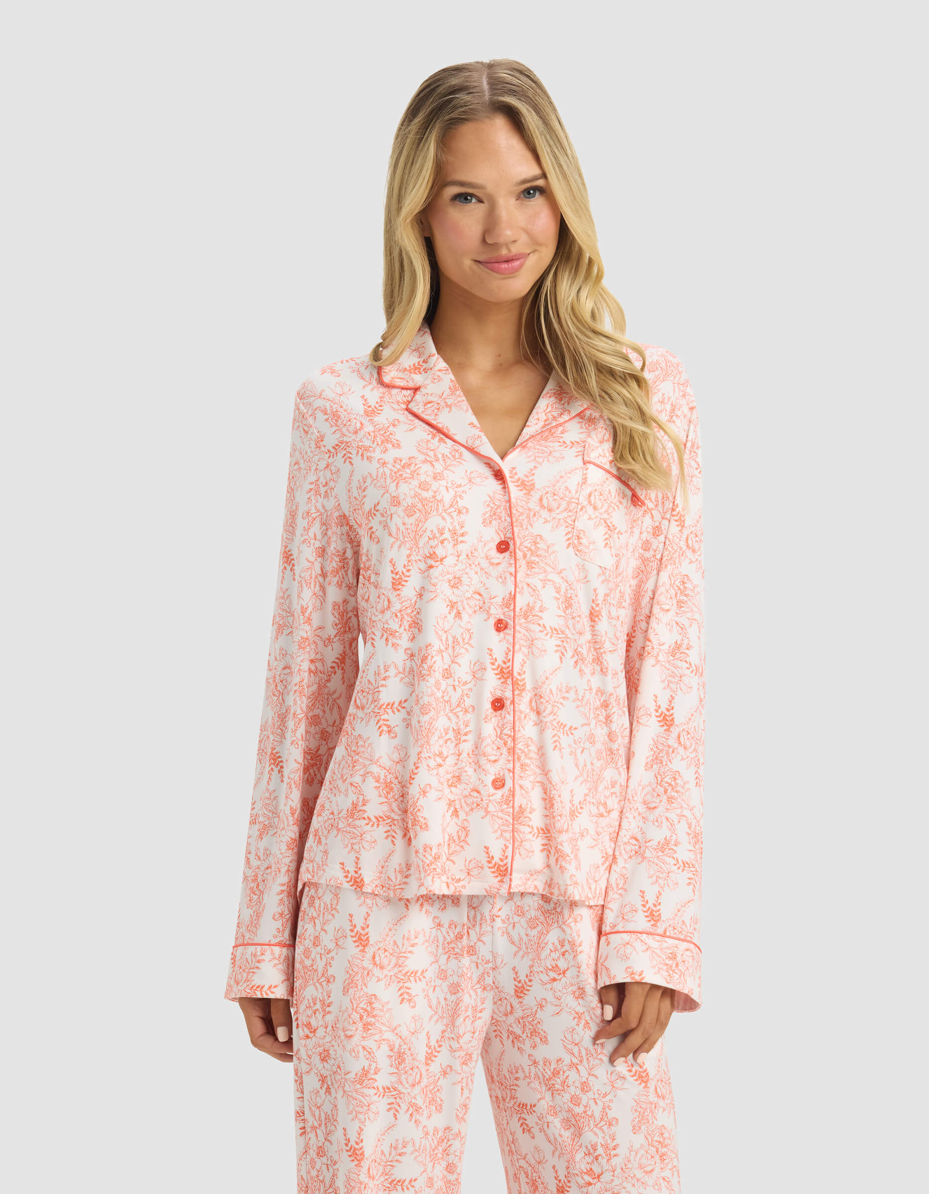 A woman with long blonde hair wears the Cozy Earth Women's Bamboo Stretch-Knit Long Sleeve Pajama Set in a light pink and white floral pattern, including a button-up top and pants, standing against a plain light gray background.