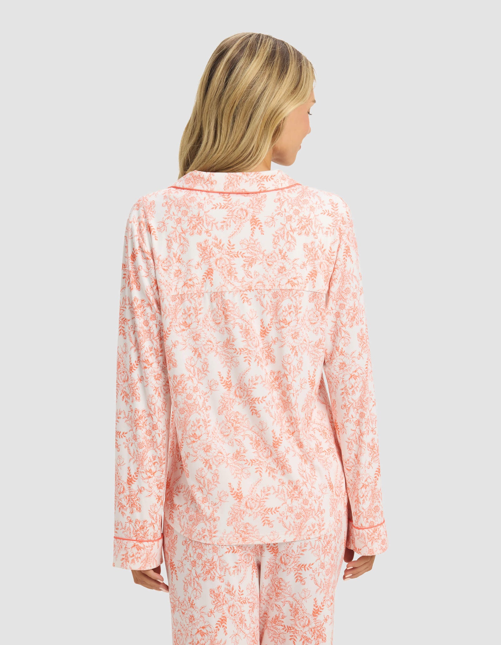 A woman with blonde hair is seen from the back wearing Cozy Earth’s Women's Bamboo Stretch-Knit Long Sleeve Pajama Top in a light pink and white floral print, paired with matching pants, against a plain light gray background.