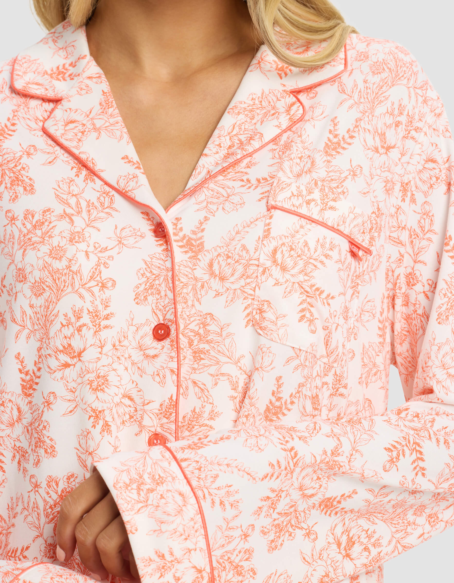 A woman wears the Cozy Earth Women's Bamboo Stretch-Knit Long Sleeve Pajama Set in light pink with a red floral pattern, red piping on the collar and cuffs, and red buttons. Her hand rests on her sleeve; her face is partially out of view.