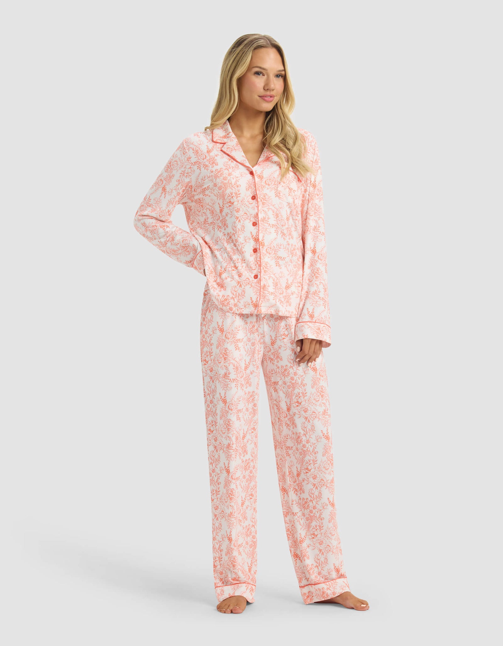 A woman stands barefoot in Cozy Earth’s Women's Bamboo Stretch-Knit Long Sleeve Pajama Set, featuring a pink and white pattern with a button-up top and matching pants, against a plain light gray background.