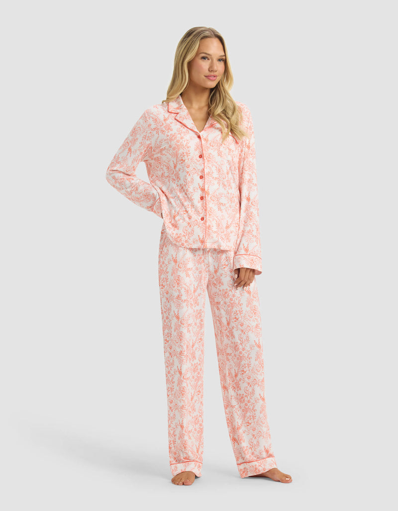 A woman stands barefoot in Cozy Earth’s Women's Bamboo Stretch-Knit Long Sleeve Pajama Set, featuring a pink and white pattern with a button-up top and matching pants, against a plain light gray background.