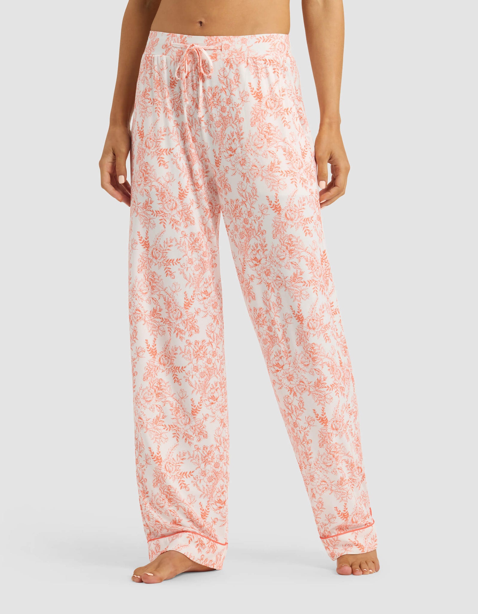 A person models the lower torso and legs wearing Cozy Earth's Women's Bamboo Stretch-Knit Long Sleeve Pajama Set in a light pink and white floral print, standing barefoot against a plain background.