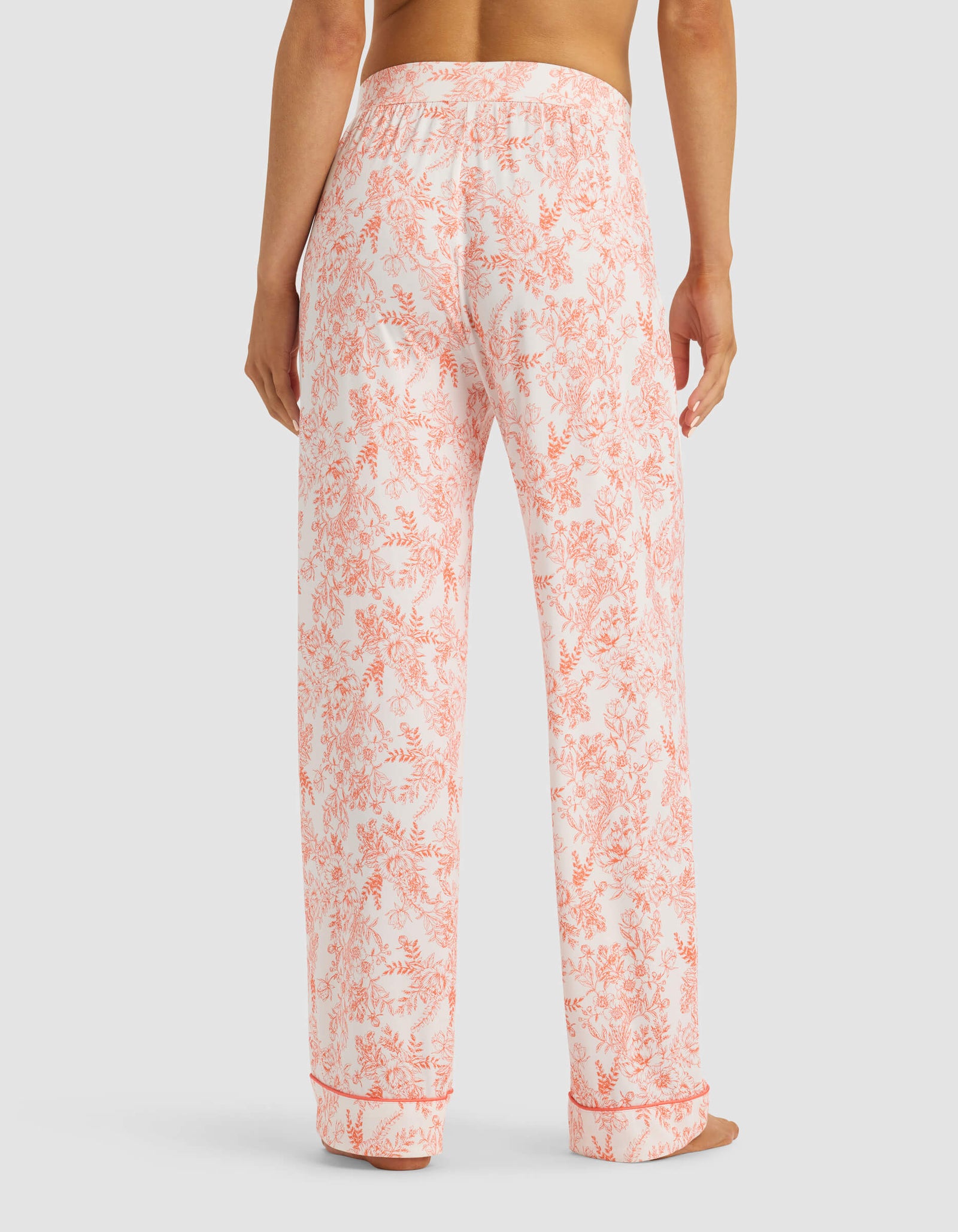A person is shown from the back wearing Cozy Earth Women's Bamboo Stretch-Knit Pajama Pant in white with a pink floral pattern. The pants have a loose, relaxed fit. The upper body of the person is not fully visible.