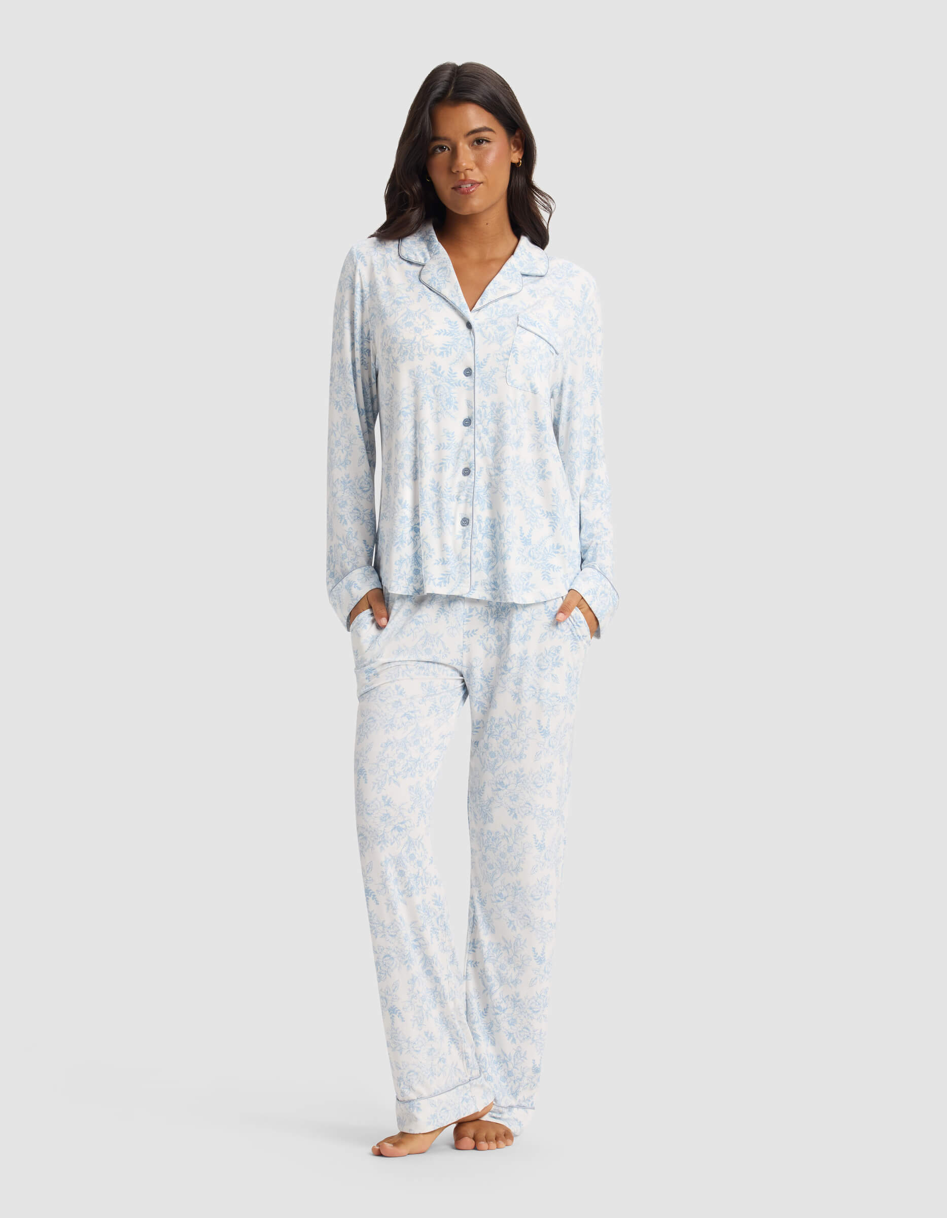 A woman stands barefoot, hands in her pockets, wearing the Cozy Earth Women's Bamboo Stretch-Knit Long Sleeve Pajama Set in a light blue and white pattern, against a plain light gray background.