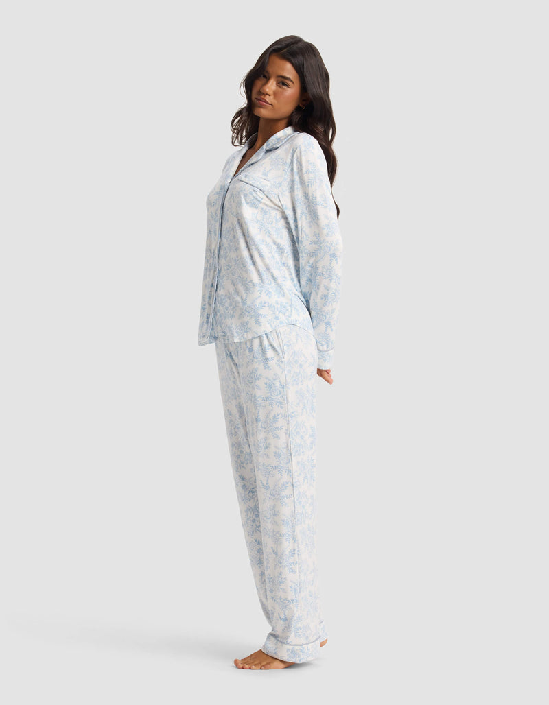 A woman models the Cozy Earth Women's Bamboo Stretch-Knit Long Sleeve Pajama Set in light blue and white, standing barefoot against a plain gray background with her arms behind her back and head slightly tilted.