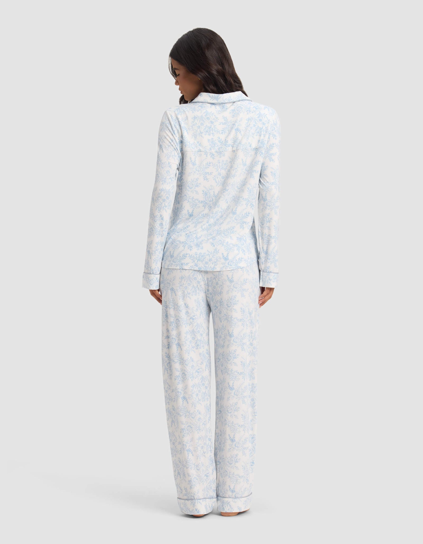 A woman faces away wearing the Cozy Earth Women's Bamboo Stretch-Knit Long Sleeve Pajama Set in light blue and white, featuring a delicate floral pattern, against a plain light background.