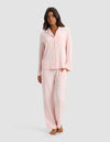 A woman stands barefoot in a Cozy Earth Women’s Bamboo Stretch-Knit Long Sleeve Pajama Set—light pink, button-up top with chest pocket and matching loose pants—against a plain light gray background.|Color:Blossom