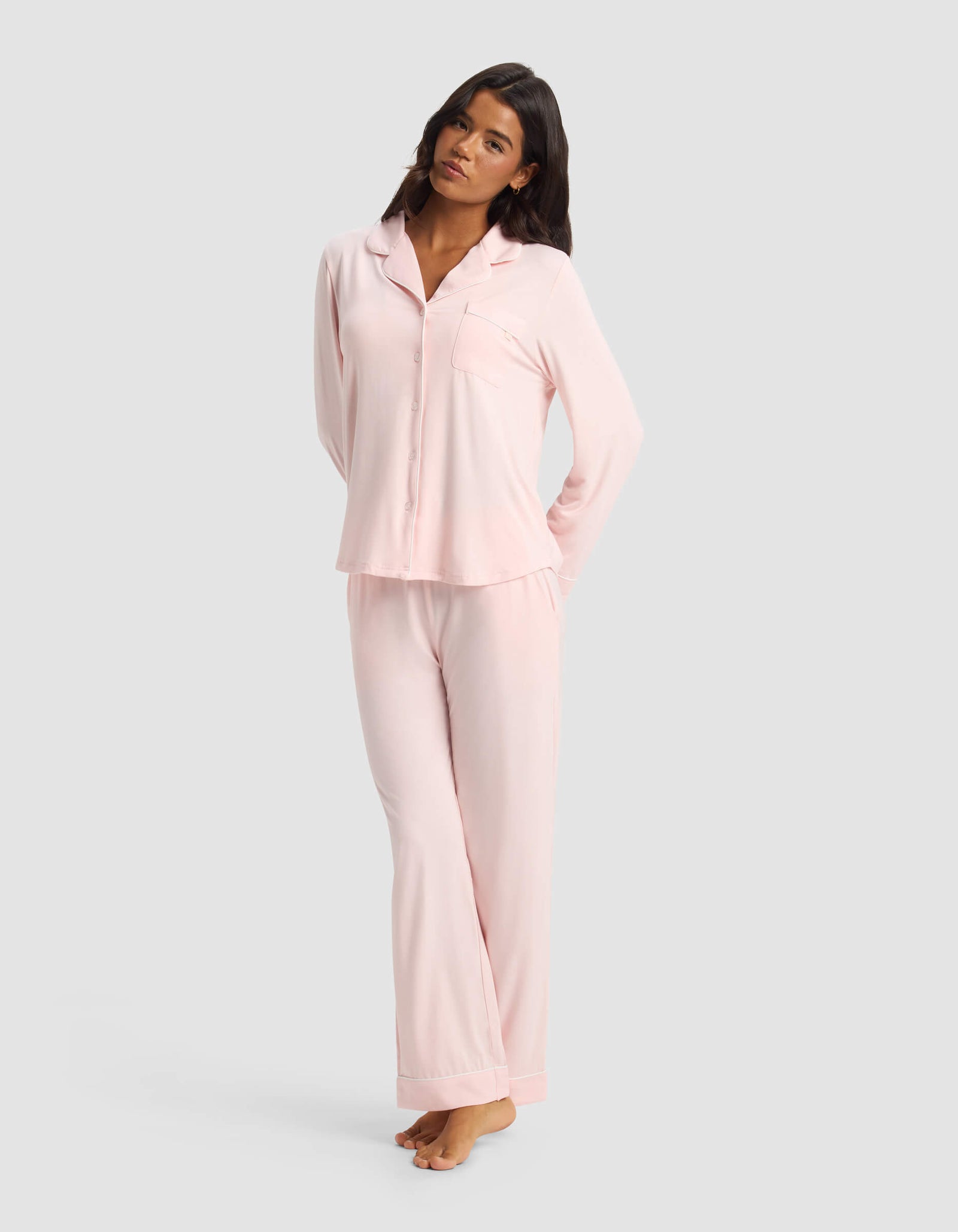 A woman with long dark hair stands barefoot against a light gray background, wearing Cozy Earth’s Women's Bamboo Stretch-Knit Long Sleeve Pajama Set—light pink, with a button-up top and matching pants. Her arms are relaxed behind her back.