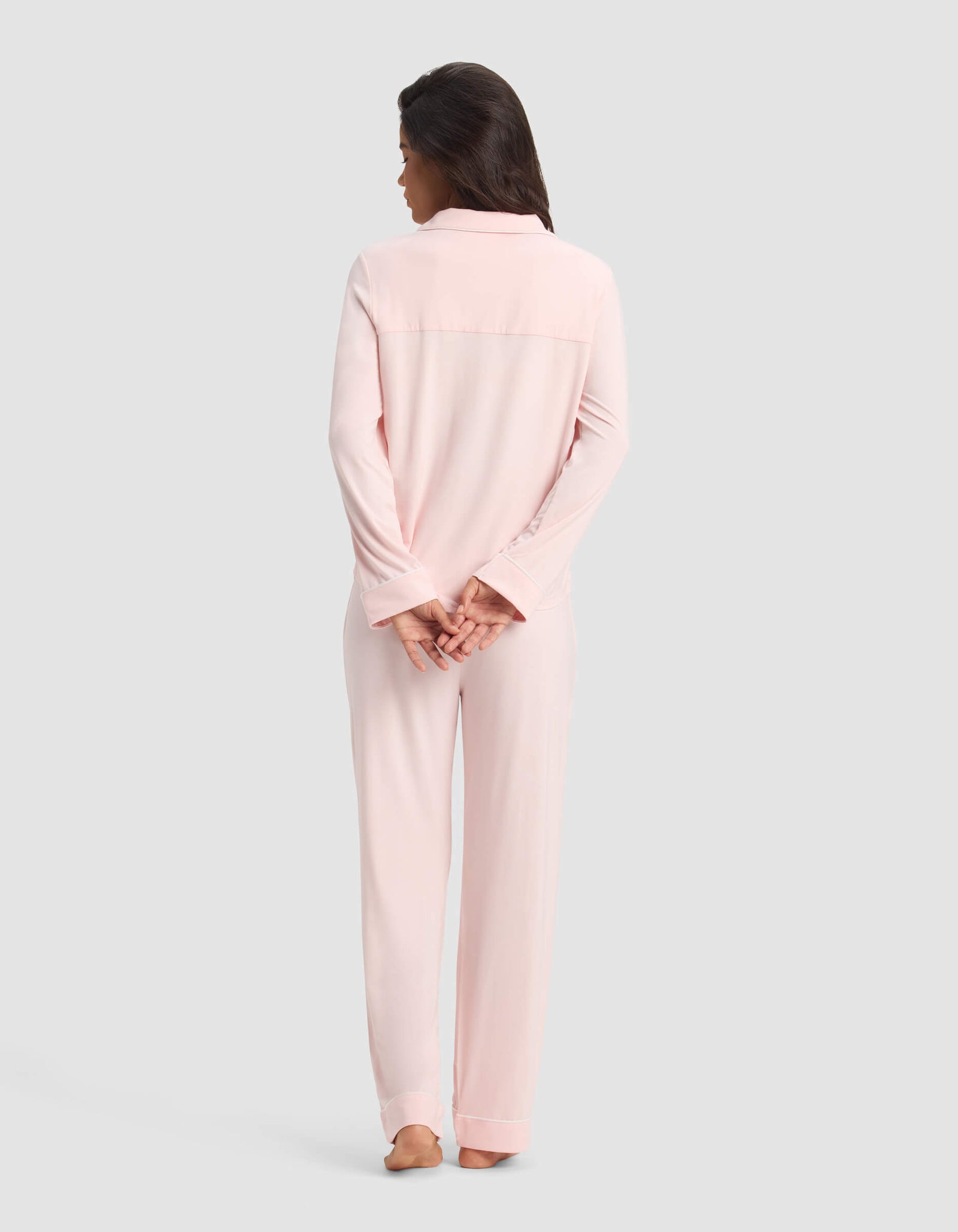 A woman stands barefoot facing away in the Cozy Earth Women's Bamboo Stretch-Knit Long Sleeve Pajama Set in light pink. Her dark hair is loose, hands clasped behind her back, against a plain light gray background.