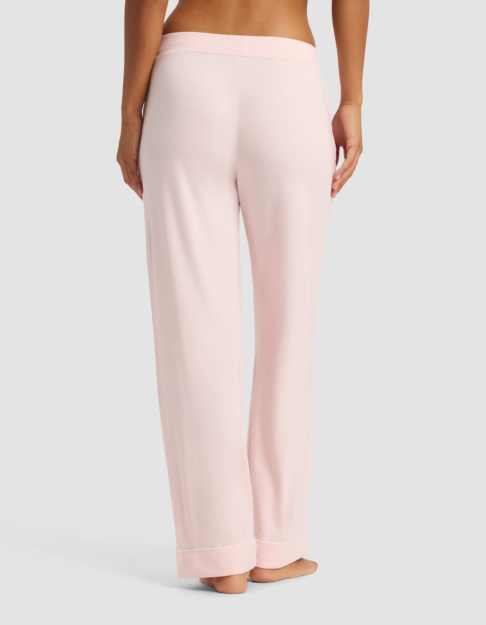 A person is standing barefoot on a neutral background, shown from the back, wearing Cozy Earth Women's Bamboo Stretch-Knit Pajama Pants in light pink with a wide waistband.|Color:Blossom