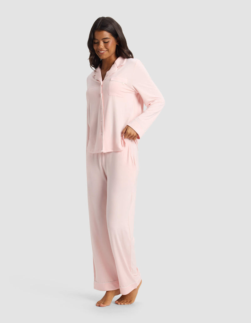 A woman stands barefoot on a white background, smiling and looking down while wearing Cozy Earth’s Women's Bamboo Stretch-Knit Pajama Pant in light pink with a matching long-sleeve button-up top.