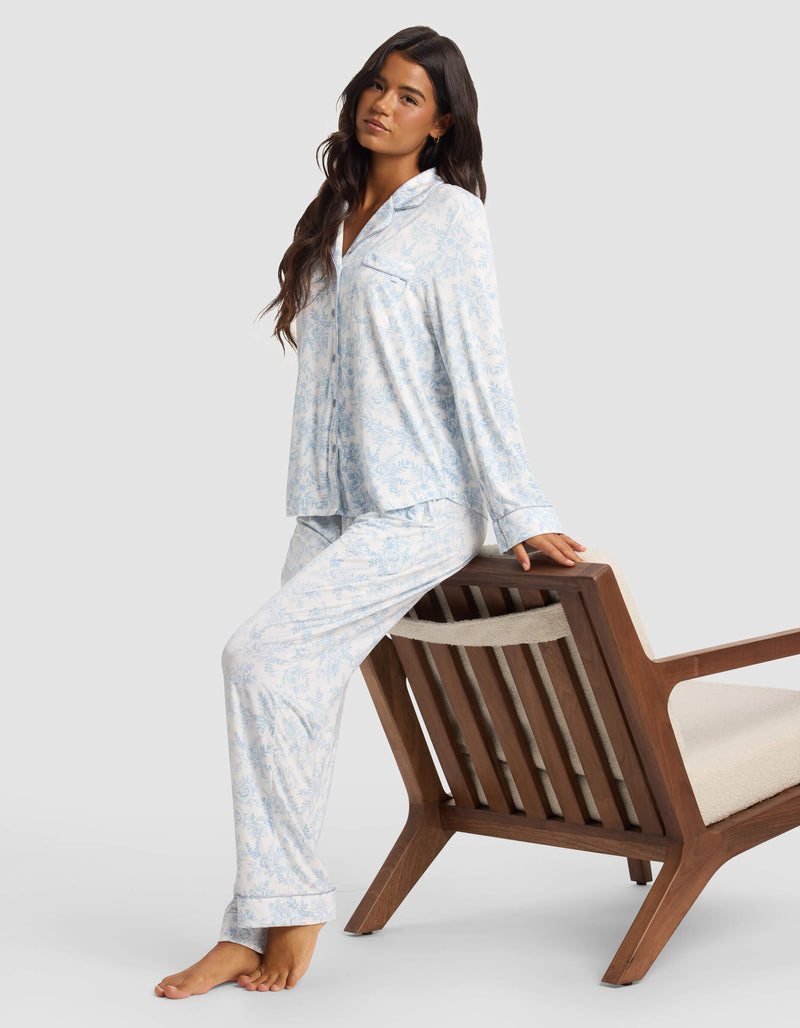 A woman with long dark hair sits barefoot on a wooden chair, wearing Cozy Earth's Women's Bamboo Stretch-Knit Pajama Pant in a light blue and white pattern, gazing calmly forward against a neutral background.
