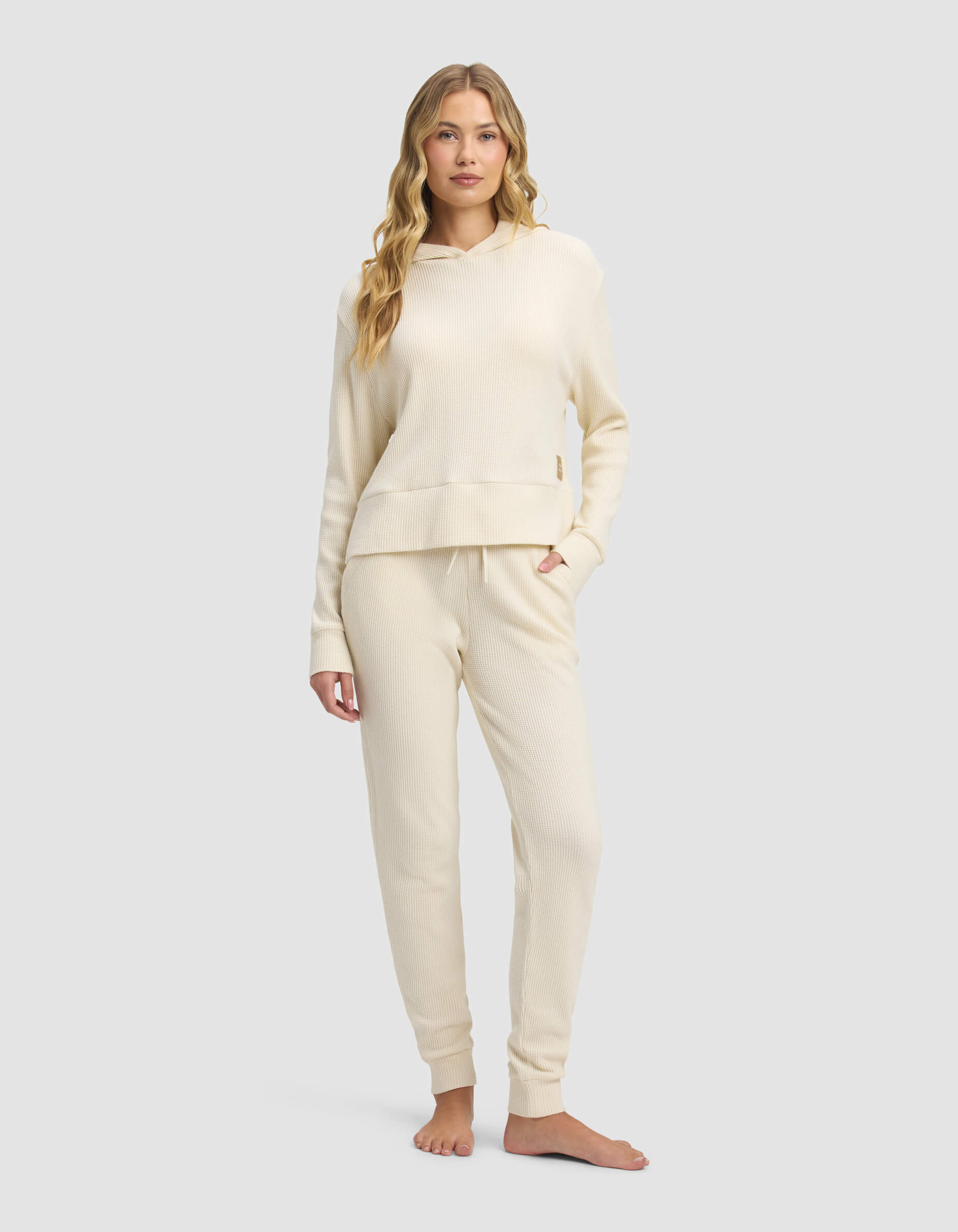 A woman stands barefoot against a light gray background, wearing the Cozy Earth Women's Blake Waffle Hoodie & Jogger Set in cream. She faces forward with one hand in her pocket.