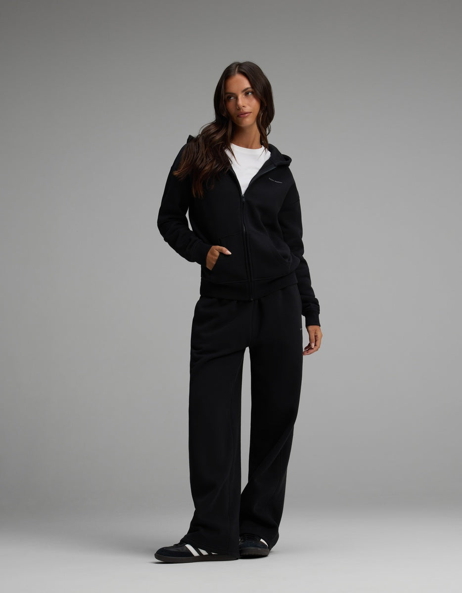 A person wearing a black Cozy Earth Women’s CityScape Full-Zip Hoodie over a white t-shirt, paired with matching black wide-leg sweatpants and black and white sneakers.
