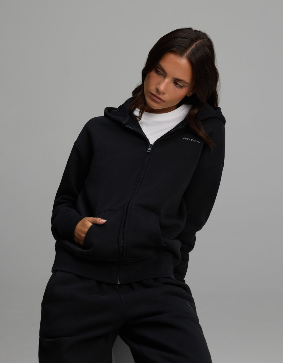 A woman wearing a black Cozy Earth Women’s CityScape Full-Zip Hoodie over a white t-shirt and matching black sweatpants.