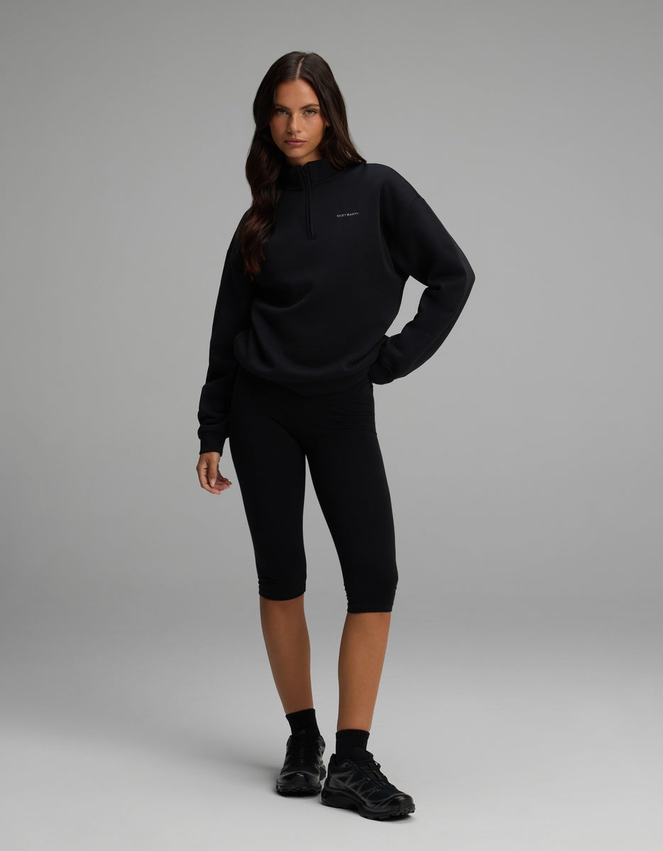 A person wearing a black Cozy Earth Women’s CityScape Quarter Zip and black capri-length leggings.