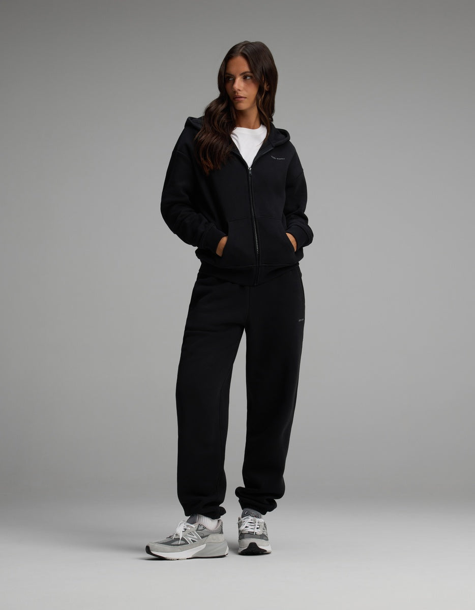 A woman wearing a black Cozy Earth Women’s Cityscape Full-Zip Hoodie & Sweatpant Set over a white shirt, paired with grey sneakers.