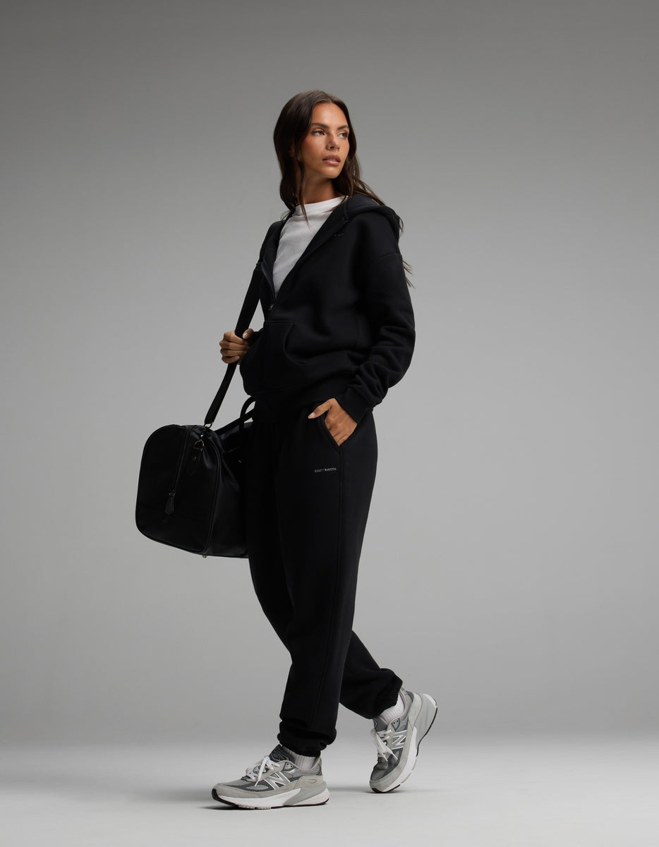 A woman wearing a black Cozy Earth Women’s Cityscape Full-Zip Hoodie & Sweatpant Set carries a black duffel bag and wears grey and white athletic sneakers.