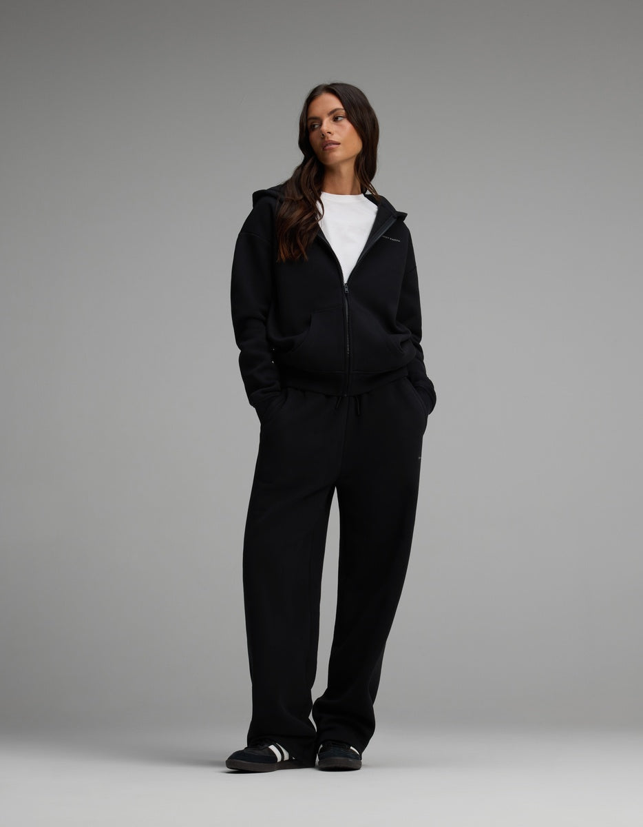 A woman wearing a black Women’s Cityscape Full-Zip Hoodie over a white t-shirt and matching black wide-leg pants.