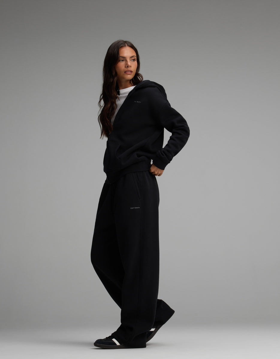 A person wearing a black Women’s Cityscape Full-Zip Hoodie over a white shirt and matching black wide-leg pants.