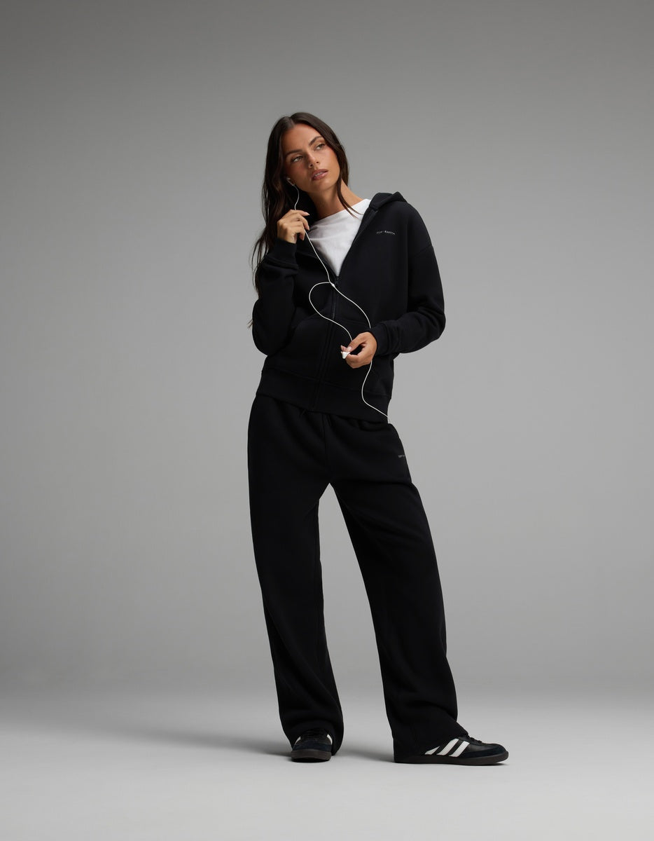 A woman wears a black Women’s Cityscape Full-Zip Hoodie & Wide Leg Pant Set over a white t-shirt, paired with sneakers.