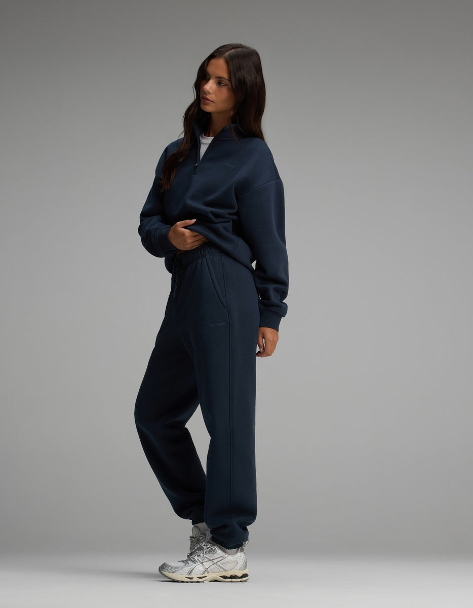 A woman wearing a dark navy blue Women’s Cityscape Quarter Zip & Sweatpant Set with matching sweatpants and metallic grey sneakers.