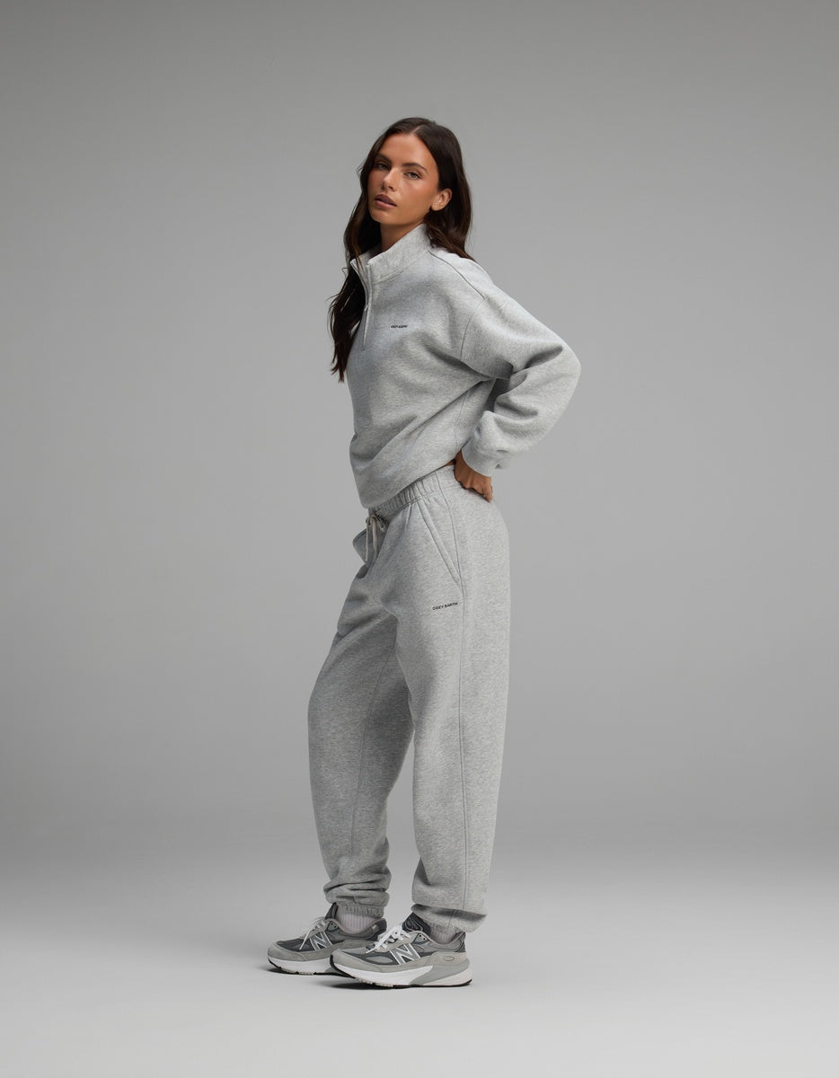 A woman standing sideways wearing a heather gray Women’s Cityscape Quarter Zip & Sweatpant Set and matching gray athletic sneakers.