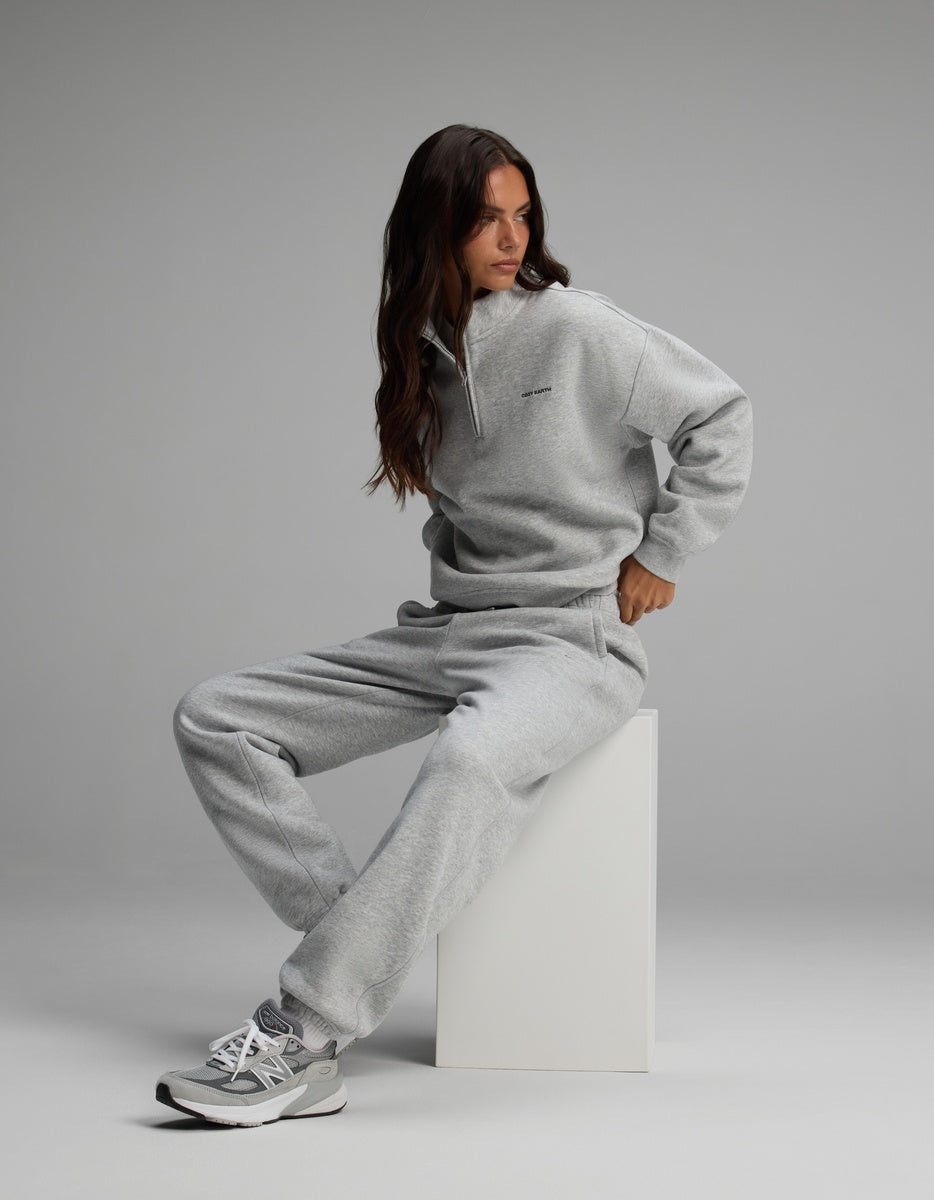 A woman wearing a grey Women’s Cityscape Quarter Zip & Sweatpant Set, sitting on a white block and wearing grey athletic sneakers.
