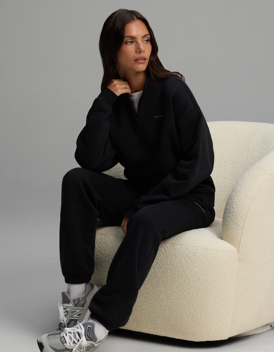 A woman wears a black Cozy Earth Women’s Cityscape Quarter Zip & Sweatpant Set while sitting in a cream-colored armchair.