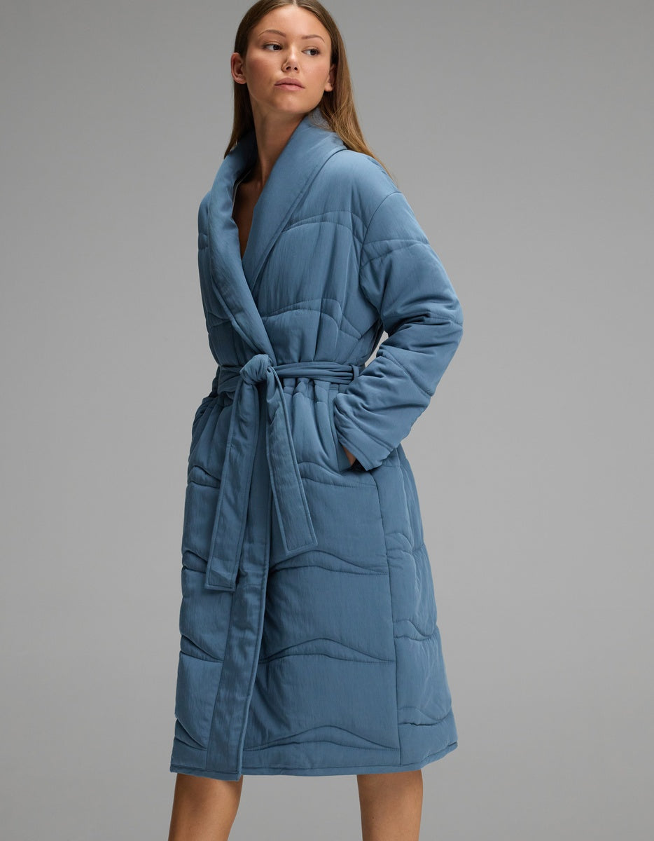 A woman wears a knee-length, quilted blue Women’s Quilted House Coat by Cozy Earth, featuring a shawl collar and a tie-waist belt.