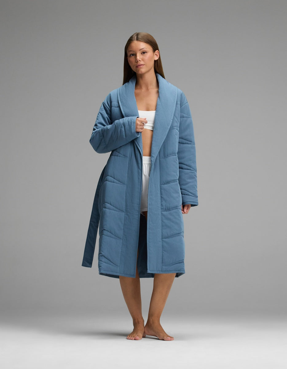 A woman wearing a blue, knee-length Cozy Earth Women’s Quilted House Coat with a wide collar and tie waist.