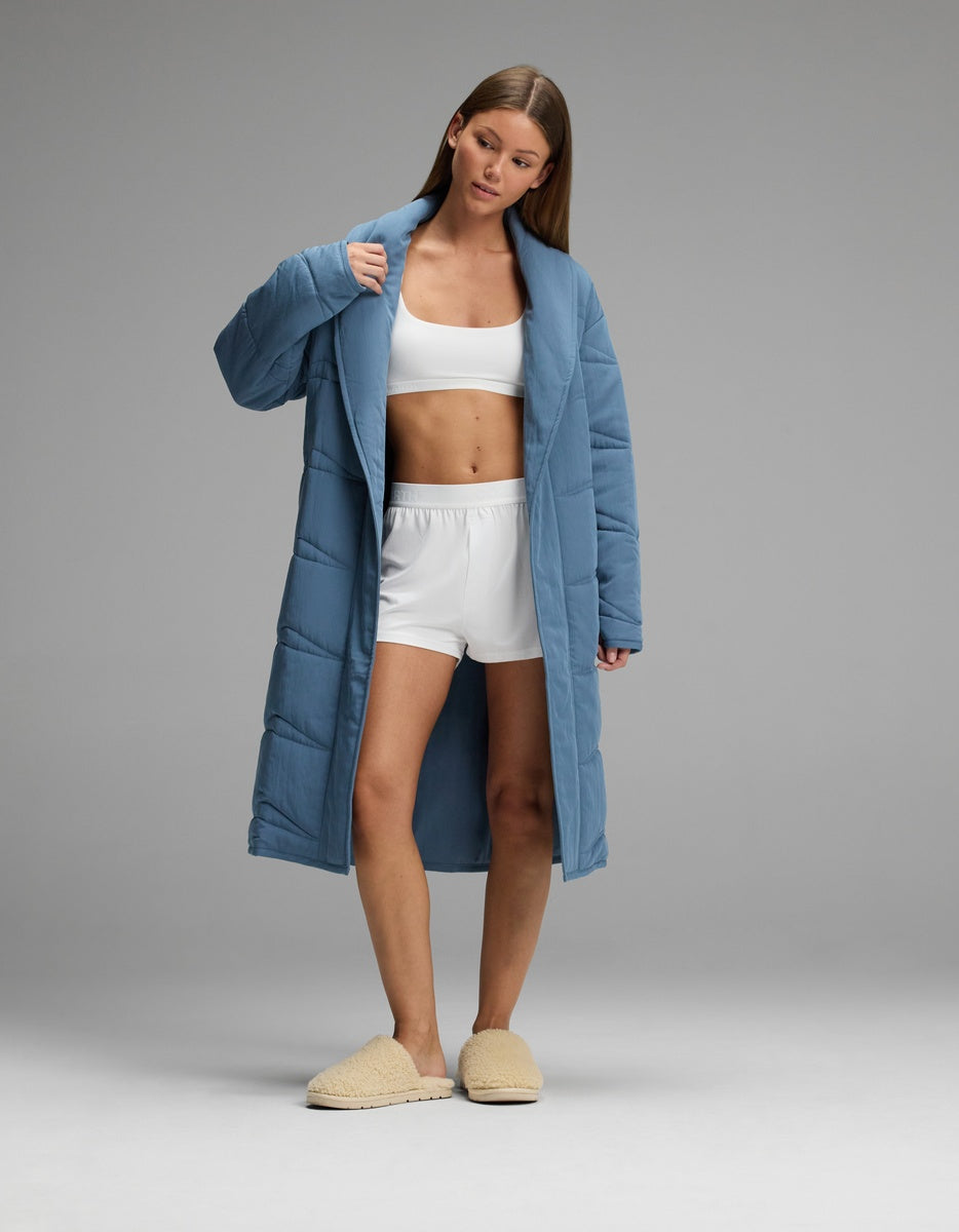 A person wearing a blue Cozy Earth Women’s Quilted House Coat over a white sports bra and shorts, with beige fuzzy slippers.