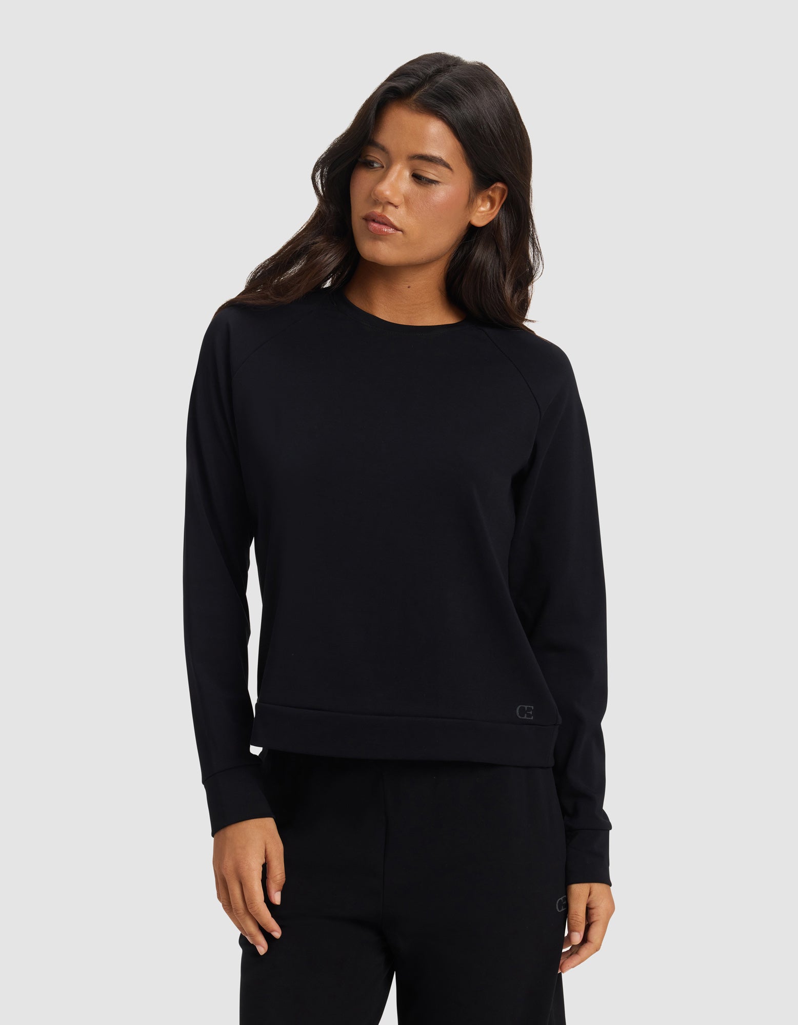 A woman with long dark hair wears the Cozy Earth Women's Brushed Bamboo Pullover Crew and matching black pants, standing against a light gray background and looking slightly to her left.