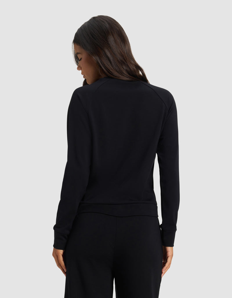 A woman with long dark hair faces away, wearing the Cozy Earth Women's Brushed Bamboo Pullover Crew in black, paired with matching black pants against a light gray background.