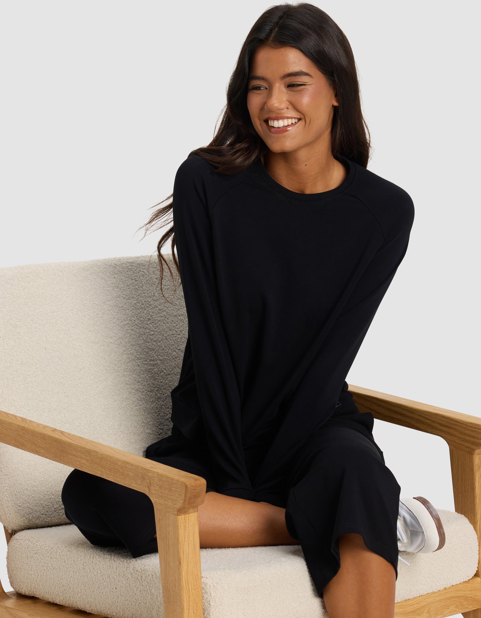A woman with long dark hair sits cross-legged, smiling, in a cream chair with wooden arms, wearing Cozy Earth's Women's Brushed Bamboo Pullover Crew in black and matching black pants.