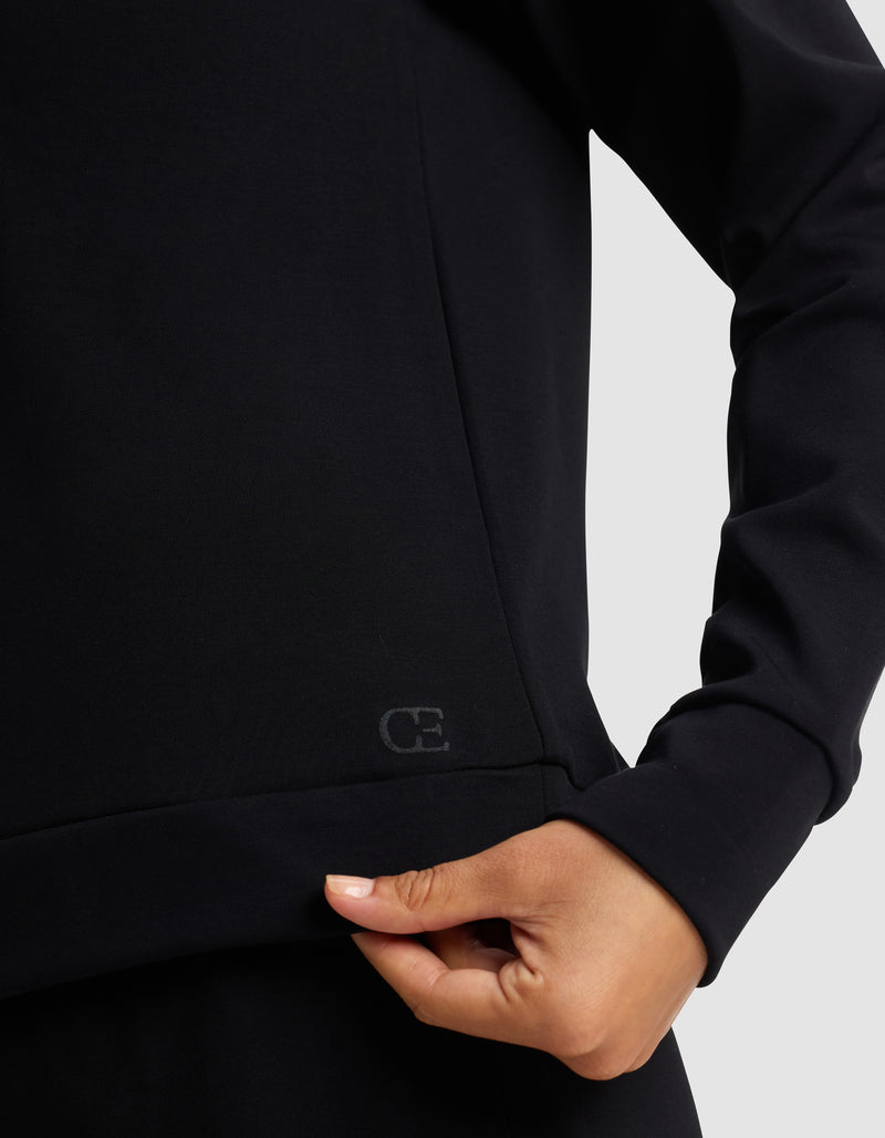 A close-up of a woman wearing the Cozy Earth Women's Brushed Bamboo Pullover Crew, gently pinching the hem with her right hand. The soft black fabric features a small, subtle "CE" logo near the bottom.