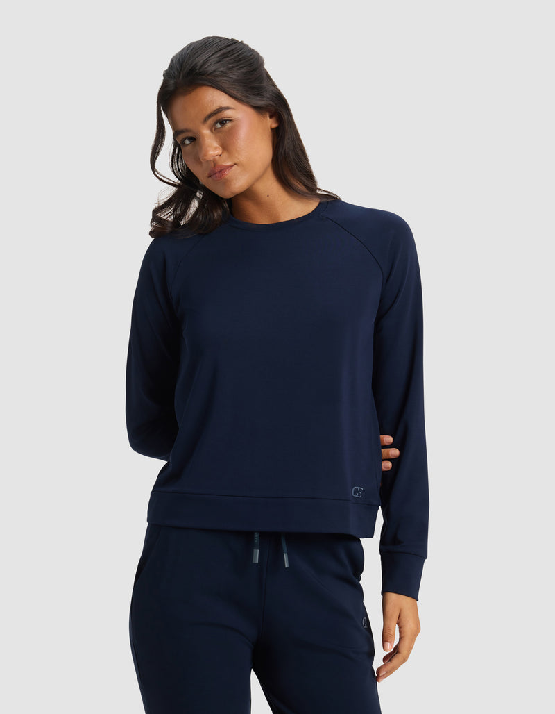 A woman with long dark hair wears the Cozy Earth Women's Brushed Bamboo Pullover Crew in navy blue, paired with matching pants, standing against a plain light gray background. She looks slightly to the side with a relaxed expression.