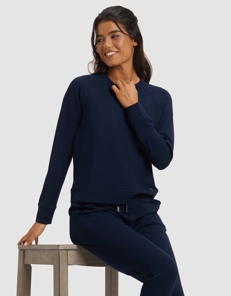 A woman in a Cozy Earth Women's Brushed Bamboo Pullover Crew sits on a wooden stool, smiling and looking to the side against a plain light background.