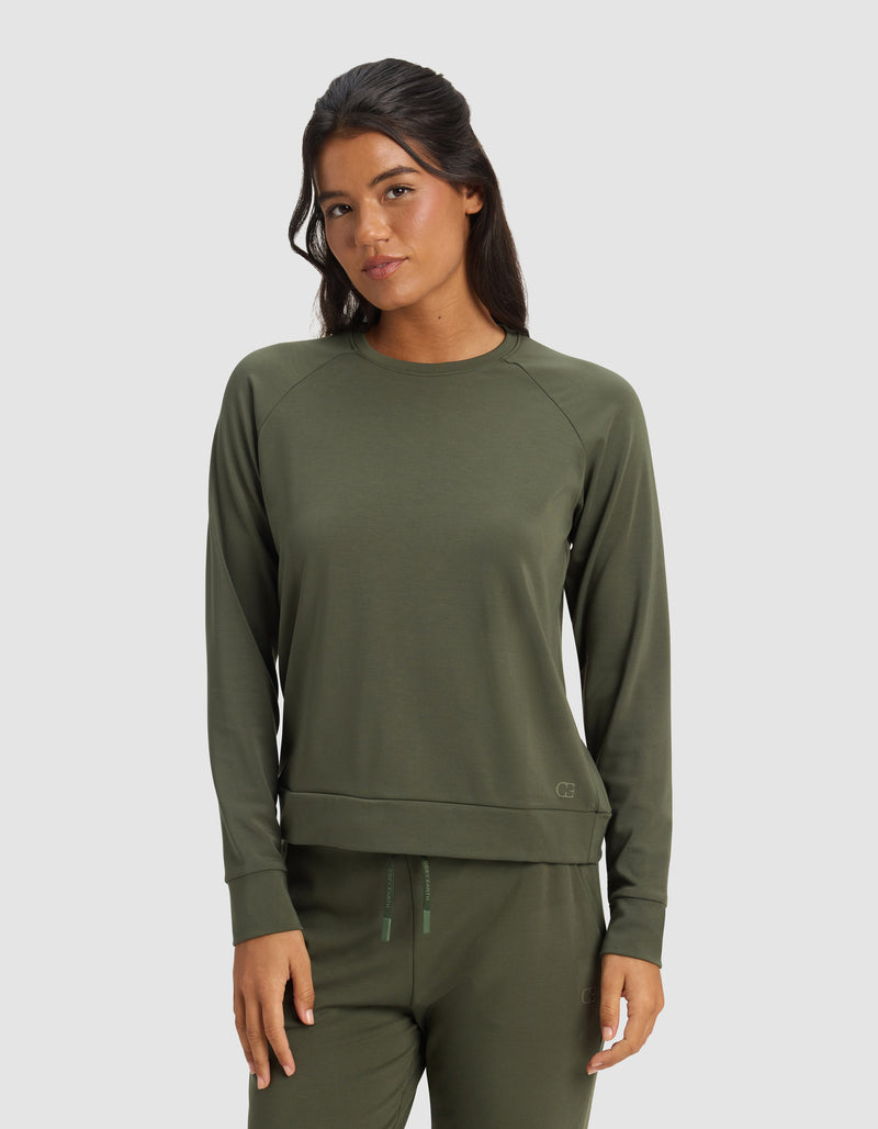 A woman with long dark hair wears the Cozy Earth Women's Brushed Bamboo Pullover Crew in olive green, paired with matching pants, standing against a plain light gray background.